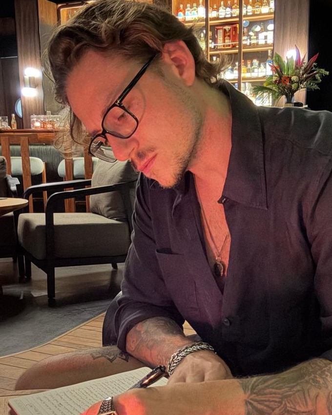 Marco, in profile, writing in a journal by candlelight with tattoos visible